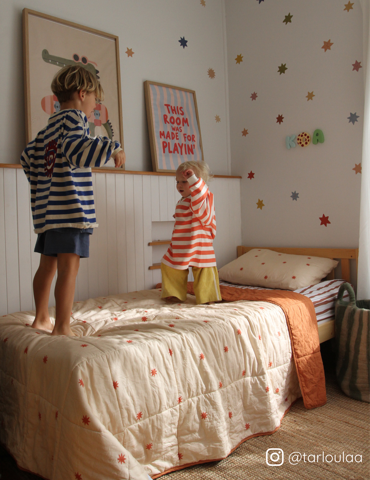 Two children jumping on a bed in a kids bedroom featuring the cuddle collective organic cotton bedding. The bed has cream bedding with red star patterns and a stripe sheet set