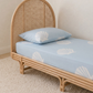 A light blue organic cotton fitted sheet and matching pillowcase with a white seashell pattern, shown on a natural rattan single bed in a softly lit room.