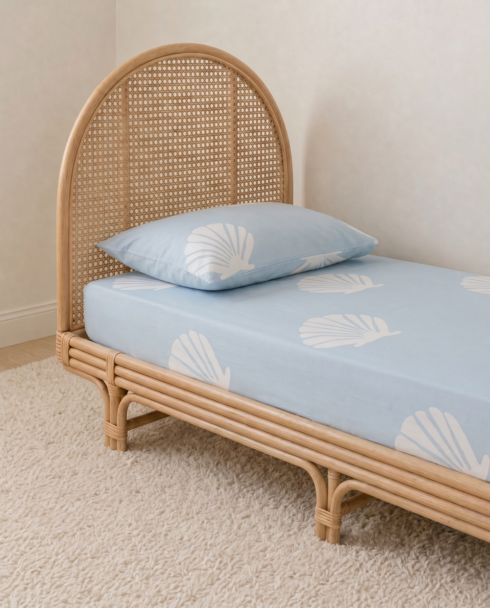 A light blue organic cotton fitted sheet and matching pillowcase with a white seashell pattern, shown on a natural rattan single bed in a softly lit room.