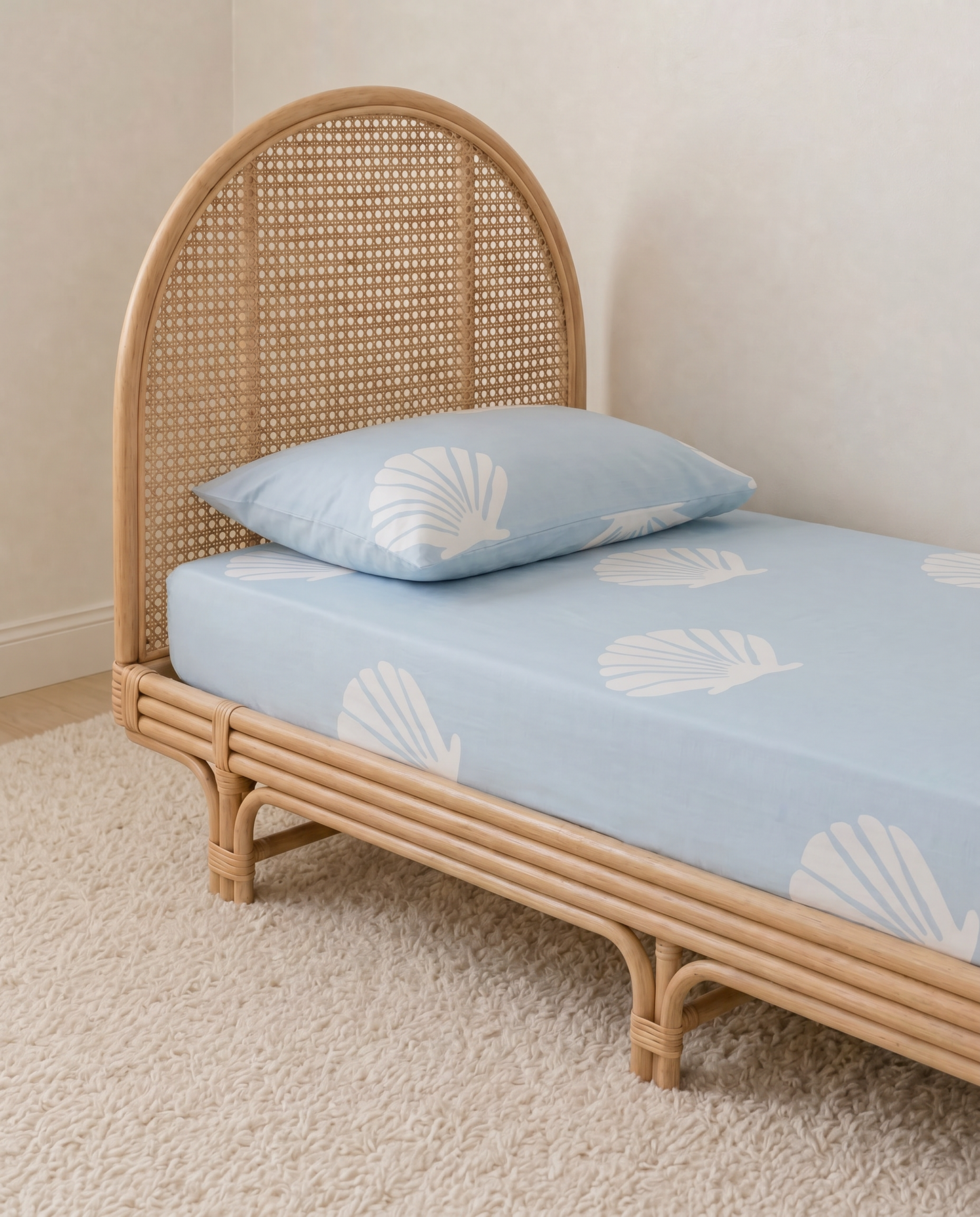 A light blue organic cotton fitted sheet and matching pillowcase with a white seashell pattern, shown on a natural rattan single bed in a softly lit room.